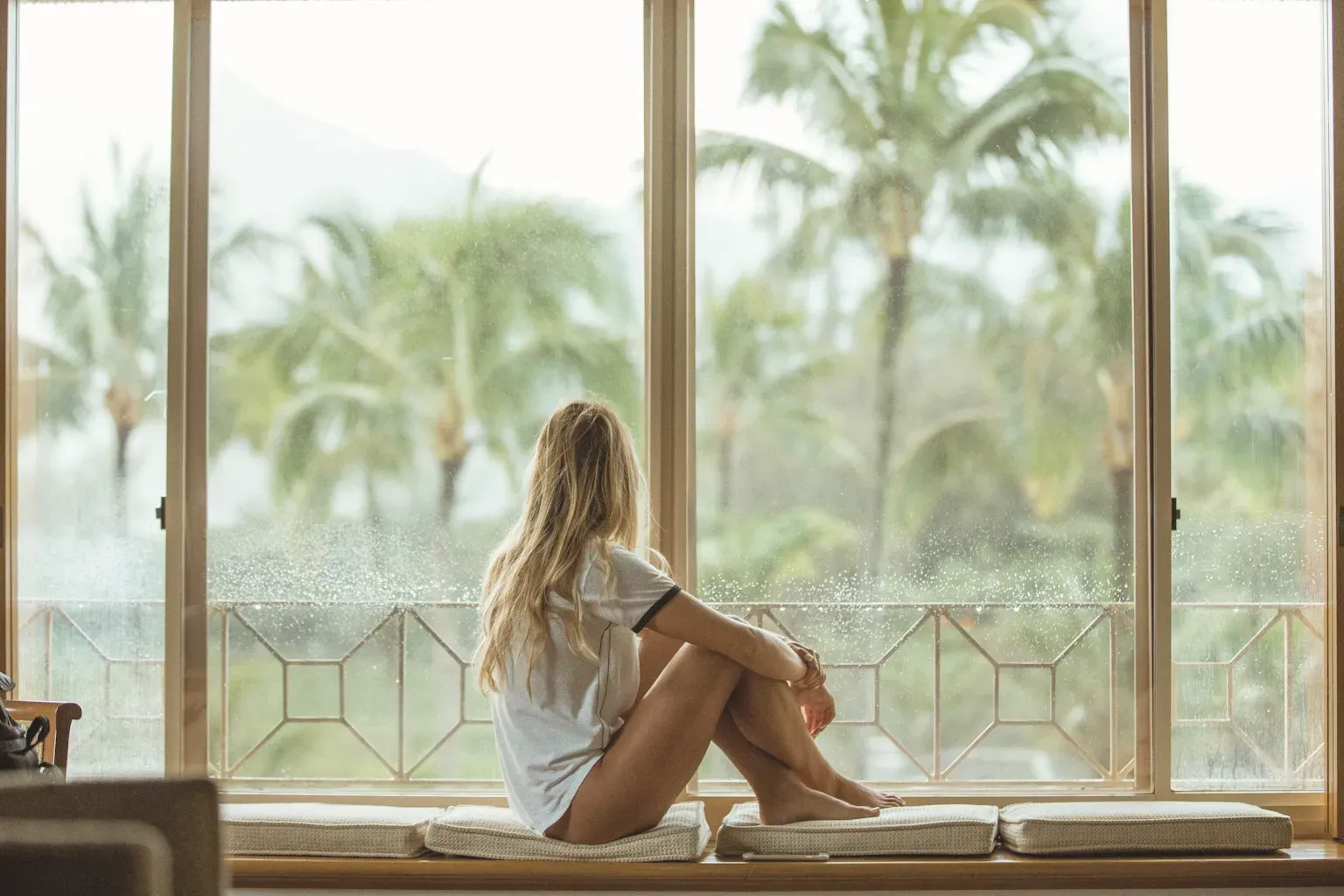 A woman sitting on a window seat, looking out at palm trees