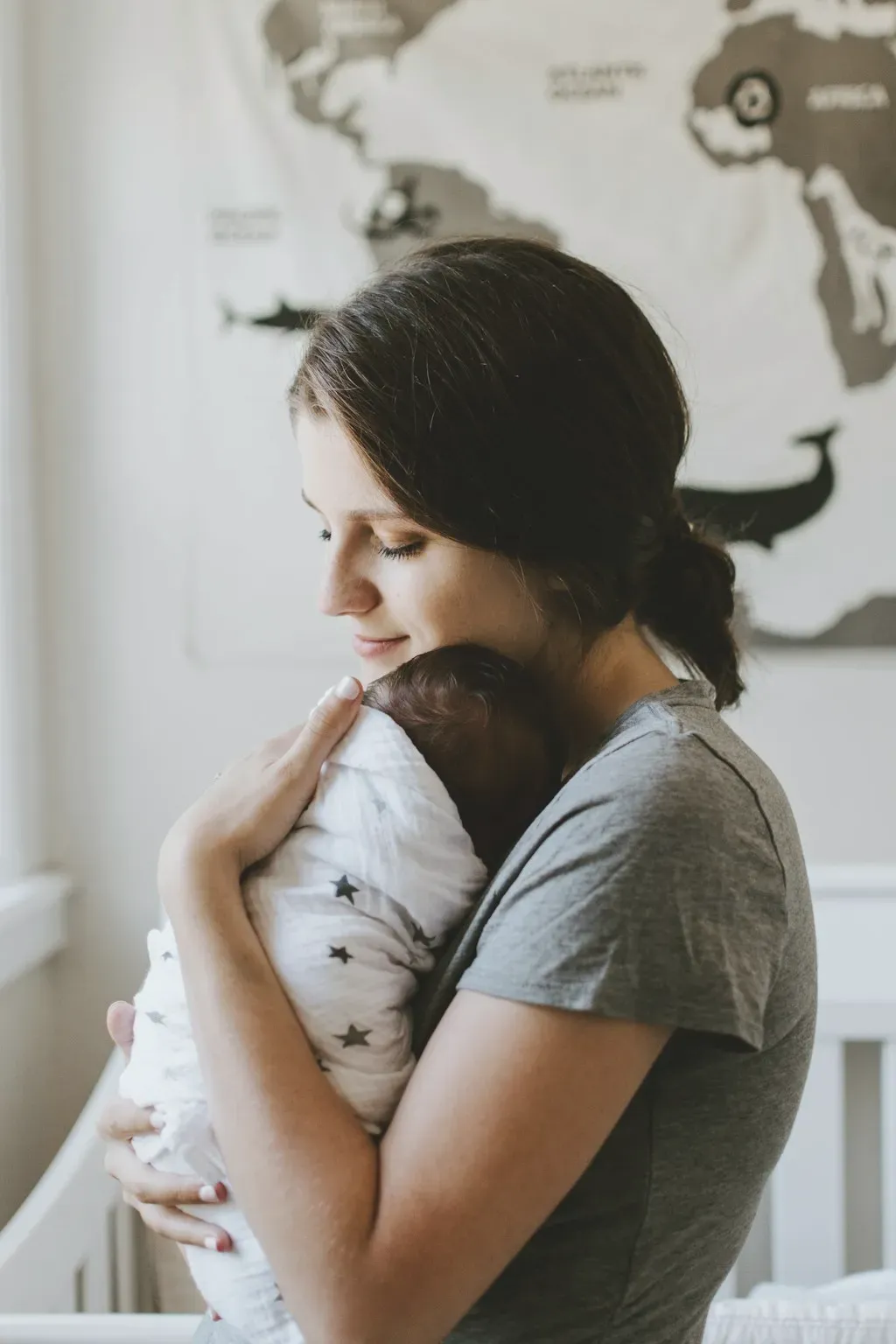 A mother holding her swaddled newborn beside a nursery wall